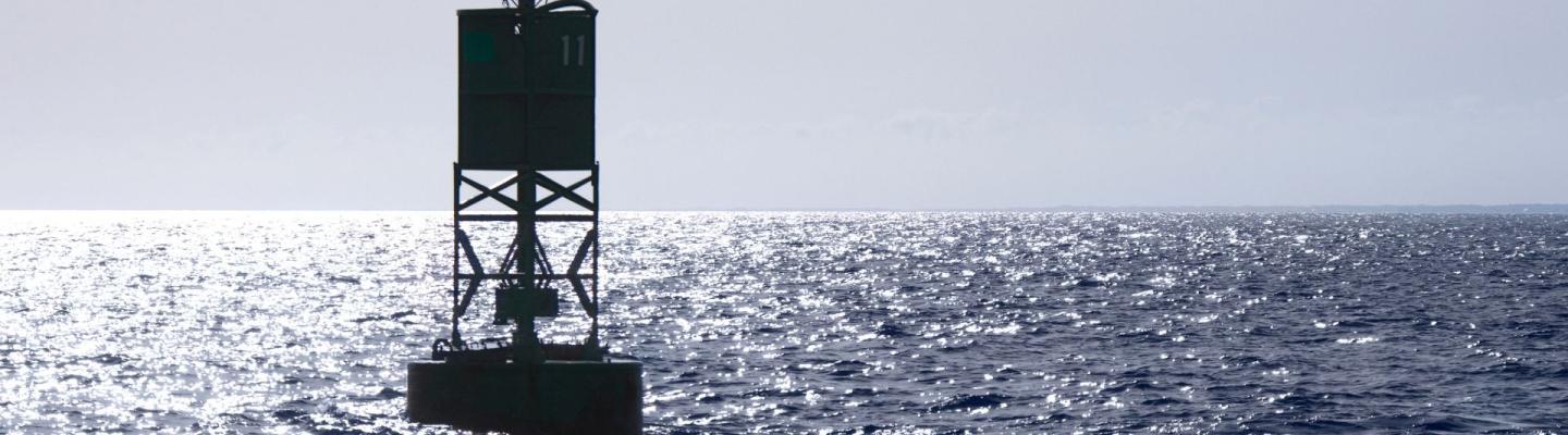 A green navigational buoy with a small light is floating in the open sea under bright sunlight and a clear sky.