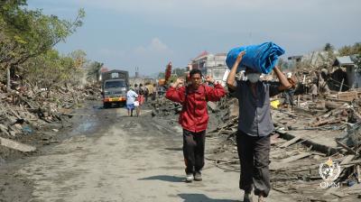 People carrying belongings walk along a debris-laden road after a disaster.