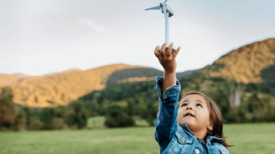 A child reaching out towards a wind turbine model in a field with hills in the background.