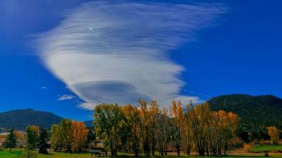 A cloud over a field with trees and mountains in the background.
