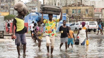 People wade through a flooded street carrying goods on their heads and in their hands in an urban setting.