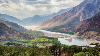 Scenic view of a winding river flowing through a valley with patchwork fields and mountain ranges under a cloudy sky.