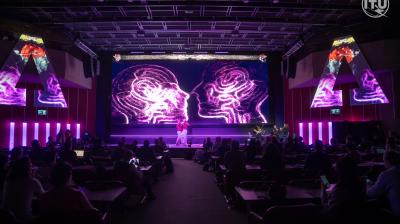 A speaker presents on a stage with a large screen displaying neon graphics of two human heads in profile. The audience is seated and attentive in a dimly lit auditorium.