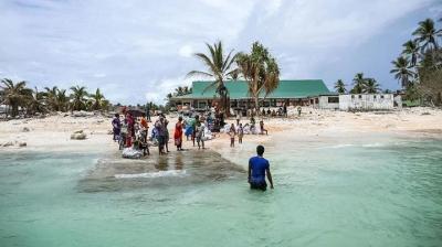 People gather on a concrete pier extending into the water from a sandy beach with palm trees and a green-roofed building in the background. One person stands in the shallow water near the pier.