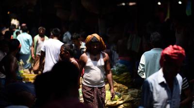 A crowded market scene with people walking and standing among various stalls in dim lighting. A man in the center wears a white tank top and a towel on his head.