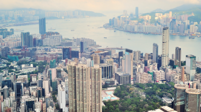 Aerial view of a dense urban cityscape with high-rise buildings, a harbor in the background, and mountains under a cloudy sky.