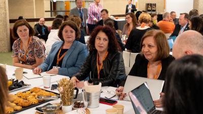 A group of people sit around a table with laptops and pastries, engaged in discussion at a conference or meeting.