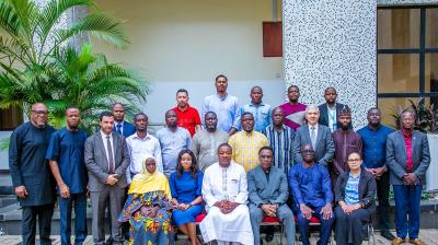 A group of people, including men and women, are posing for a photo in front of a building with a patterned façade. Some people are seated while others are standing behind them.