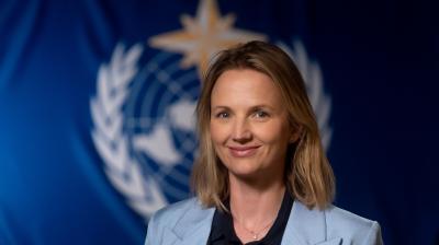 A person with shoulder-length blonde hair wearing a light blue blazer smiles in front of a blurred background featuring a blue flag with a white emblem.