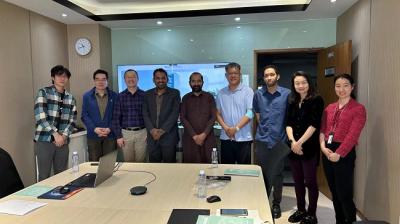 A group of eight people standing and posing for a photo in a conference room with a table, a laptop, and documents.