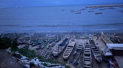 A beach with numerous wooden fishing boats lined up on the shore. Additional boats are seen floating in the water. Some structures and greenery are visible in the foreground.