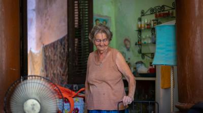 An elderly woman stands in a room with a fan, colorful cloth, and household items. She wears a sleeveless top and glasses, looking down with a gentle smile.