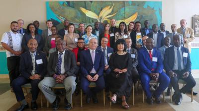 A group of 28 people, dressed in business and formal attire, pose together in front of a large, colorful wall painting for a group photo, likely at a conference or formal event.