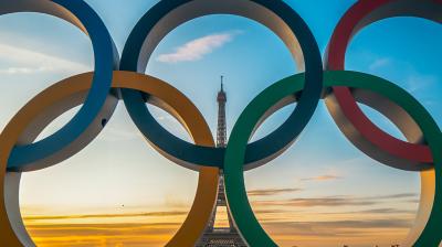 The Eiffel Tower is framed through the Olympic rings in Paris at sunset, with a colorful sky in the background.