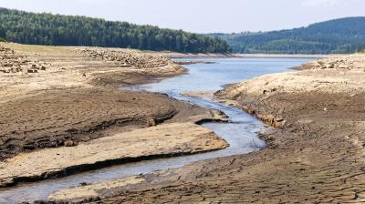 A narrow stream flows through parched, cracked earth with forests and hills in the background under a partly cloudy sky.