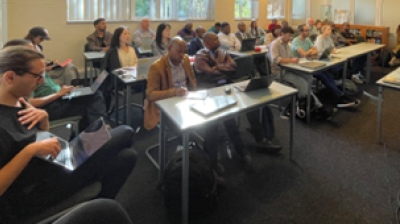 A group of people seated in a classroom, each with laptops or notepads, attentively listening to a presentation.