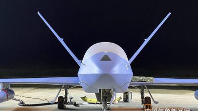 A large drone is positioned on a tarmac at night, illuminated by artificial light. Two wings are visible, along with the ground equipment and cables connected to it. Chinese text is displayed at the bottom right.