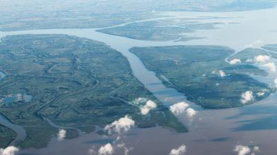 Aerial view of a river delta with multiple branching waterways and green landmasses, partially obscured by scattered clouds.