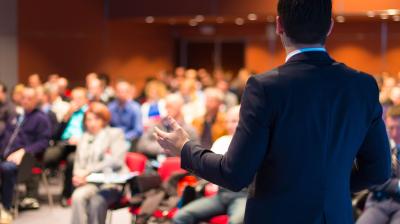 A person in a suit stands with their back to the camera, addressing a seated audience in a large, brightly lit room.