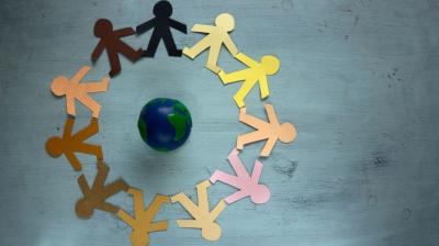 A ring of multicolored paper cutouts of people holding hands encircles a small globe on a gray background.