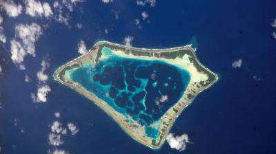 Aerial view of a triangular atoll surrounded by deep blue ocean, with a mix of shallow turquoise waters, reefs, and scattered clouds.