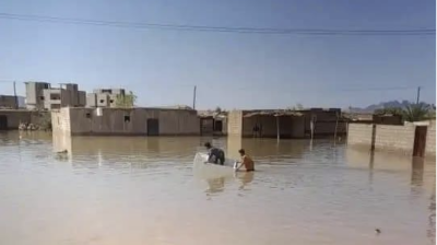 Three people in a small boat navigate through a flooded area with partially submerged buildings under a clear blue sky.