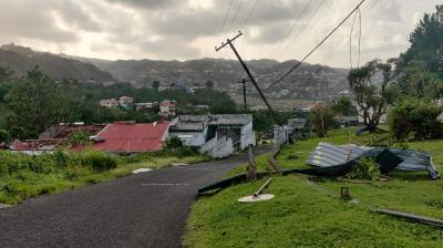 A damaged utility pole leans over a road, with debris and roofing materials scattered around in a hilly residential area. Houses and trees are visible in the background under a cloudy sky.