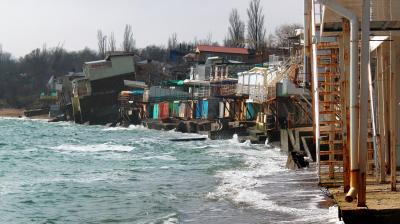 Beachfront buildings extensively damaged by erosion with portions collapsed into the sea, and waves lapping against the remains of the structures. Trees are visible in the background.