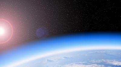 Image of Earth’s curved horizon as seen from space with the sun shining in the upper left and stars visible in the dark sky above. The planet's atmosphere and some cloud formations are also visible.