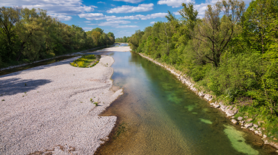 A clear, shallow river flows between gravel banks and dense green trees under a blue sky with scattered clouds.