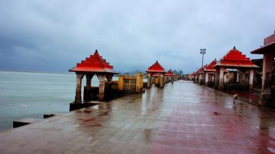A riverside promenade with red-roofed pavilions on a rainy day. The ground is wet, and the sky is overcast. The walkway is empty except for a lone dog.