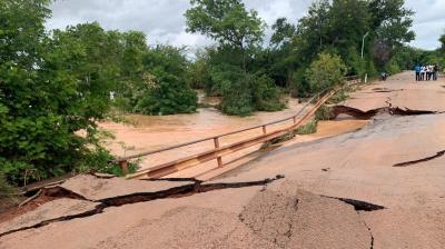A damaged road with severe cracks and a collapsed section due to flooding; muddy water flows beneath, surrounded by trees.