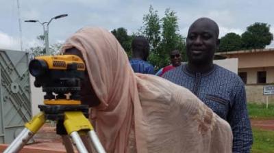 A person in a light scarf looks through a surveyor's level on a tripod outside, guided by another person standing nearby.