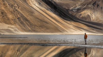 A person stands near a tripod by a reflective mountain lake with snow-covered peaks in the background.