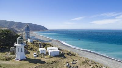 Coastal landscape featuring a white lighthouse, small buildings, a vehicle, and a tall blue structure near a beach with clear blue water and distant hills under a clear sky.