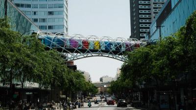 A street view showing a colorful pedestrian bridge between two tall buildings, with trees lining the road and vehicles in the distance.