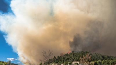 Thick smoke billows from a forest fire near a town, with red flames visible among the trees under a clear blue sky.