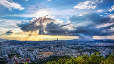Panoramic view of a cityscape with dramatic clouds and sun rays breaking through. A rainstorm is visible in the distance, surrounded by green hills and urban buildings under a blue sky.