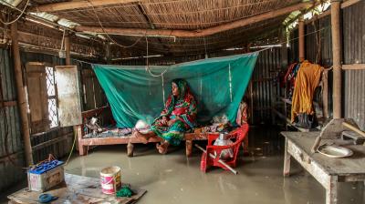 A woman sits on a bed under a mosquito net in a flood-affected home with water covering the floor. Various items, including a chair and table, are partially submerged.