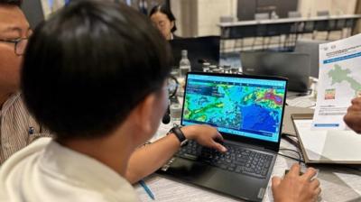 Person studying a colorful map on a laptop screen in an office setting, with papers and colleagues nearby.