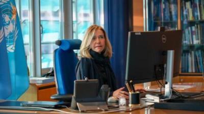 A person sitting at a desk, working on a computer in an office. A United Nations flag and bookshelf are visible in the background.
