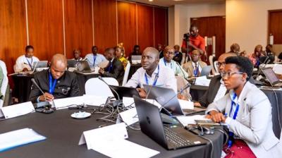 A group of people attending a conference in a meeting room, seated at round tables with laptops and notepads.