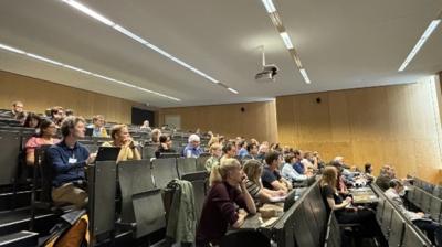 A lecture hall with attendees seated in rows, facing the front. The room has wooden walls and overhead lighting.