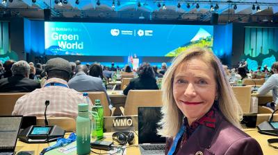 A woman in a conference hall sits at a desk with various electronic devices. A large screen displays "In Solidarity for a Green World" in the background.