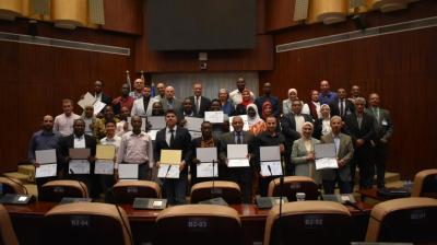 A group of people in formal attire stand in rows holding certificates inside a conference room with wooden walls and numbered seats.