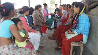 A group of people sit in a circle outdoors, some holding notebooks, engaged in discussion. A child is seated on one person's lap.