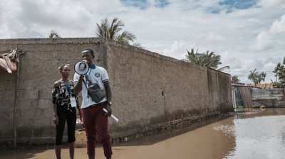 A person stands in floodwater using a megaphone, while another individual stands nearby. A partially flooded building is in the background under a cloudy sky.