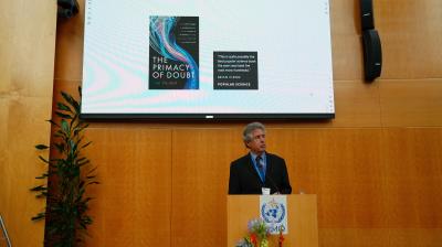 A man stands at a podium giving a presentation in front of a large screen displaying the book cover of "The Primacy of Doubt" by Tim Palmer.