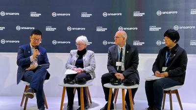 Four individuals are seated on stage, engaging in a panel discussion. Behind them is a backdrop with logos and text. Microphones and headphones are placed on the small tables in front of them.
