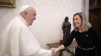 A man in a white robe and skullcap shakes hands with a smiling woman in a black dress in a room with a statue and ornate wallpaper.
